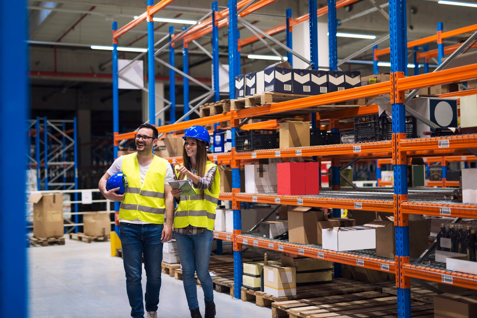 Group of warehouse workers wearing hardhats and reflective jackets waking in aisle between tall racks with packages and goods.